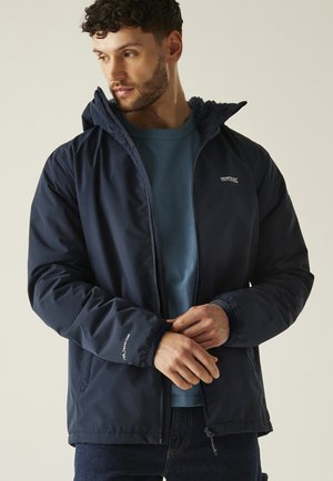 Mężczyzna w granatowej kurtce na zamek z polarem w środku i ciemnoniebieskiej koszuli, poprawiający mankiet rękawa, zwrócony lekko w lewo na jednolitym tle.