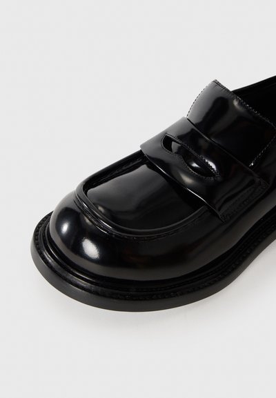 Svarta lackskinnssneakers med fyrkantig tå, ett dekorativt rem över framsidan och en något upphöjd, rund klack.