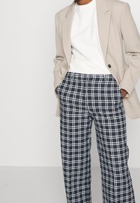 Beige kavaj över en vit t-shirt, kombinerad med navy och vit rutiga byxor. Har en avslappnad passform med framfickor och synlig sömnad.