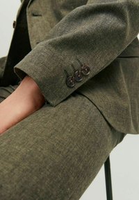 Costume vert olive en tissu texturé. Gros plan sur le poignet montrant quatre boutons foncés et des détails de coupe ajustée.