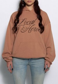 Brązowy hoodie z przednią kieszenią i szerokimi rękawami, z napisem "Local Heroes" w ciemnobrązowym kursywie. Noszony z jasnoniebieskimi dżinsami.