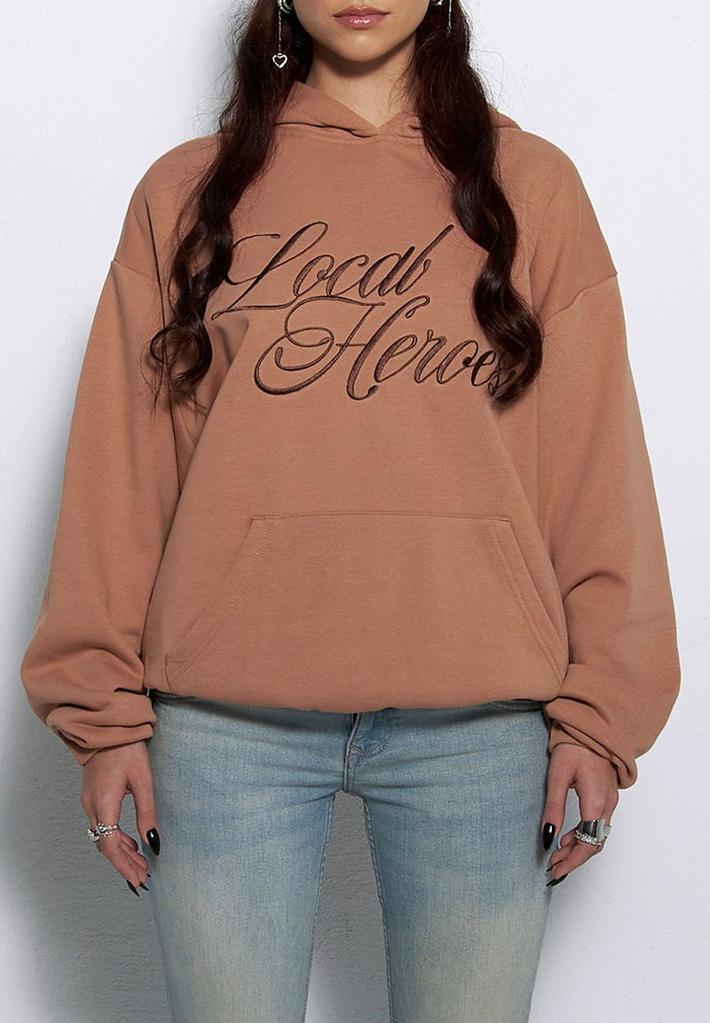 Brązowy hoodie z przednią kieszenią i szerokimi rękawami, z napisem "Local Heroes" w ciemnobrązowym kursywie. Noszony z jasnoniebieskimi dżinsami.