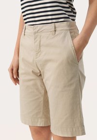 Pantaloni corti in cotone beige con davanti piatto, passanti per cintura, tasche laterali e orli arrotolati, abbinati a una maglietta a righe bianco e nera.