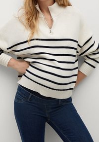 Mujer vistiendo un suéter blanco de canalé con rayas horizontales negras y un cuello con cremallera hasta un cuarto, combinado con jeans de color azul oscuro, con la mano en la cadera.