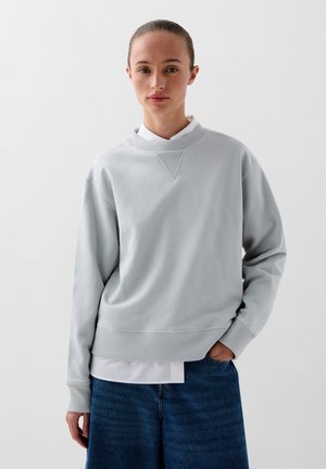 Lichtgrijze sweatshirt met een geribbelde structuur, ronde halslijn en dropped shoulders, laag over een witte blouse met kraag, gecombineerd met denim.