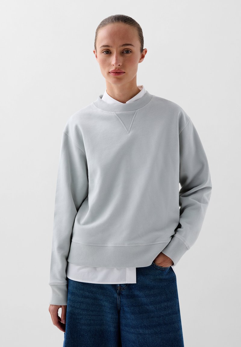 Lichtgrijze sweatshirt met een geribbelde structuur, ronde halslijn en dropped shoulders, laag over een witte blouse met kraag, gecombineerd met denim.
