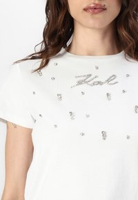 Camiseta de algodón blanca con strass dispersos y un logo "Karl" brillante en un estilo de escritura cursiva en el centro. Mangas cortas, textura suave.