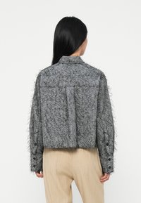 Vrouw met lang zwart haar, gekleed in een grijs gemêleerd kort jasje en lichtbeige broek, van achteren gefotografeerd tegen een effen witte achtergrond.
