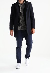 Hombre vestido con una camisa de estampado, abrigo azul marino, pantalones azul marino y zapatillas blancas, caminando frente a un fondo blanco.