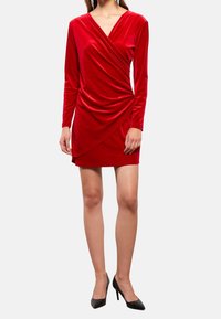Robe portefeuille en velours rouge avec des manches longues, présentant une texture drapée et une coupe courte. Associée à des escarpins noirs à talons aiguilles.
