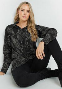 Schwarze Hemd mit Knopfleiste und abstrakten weißen Mustern, langen Ärmeln und spitzem Kragen; kombiniert mit schwarzen, eng anliegenden Hosen und Stiefeletten.