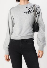 Grå sweatshirt med pufærmer, sort bladmønster broderet på skulderen, parret med sorte jeans og en lille sort taske med kædestrop.