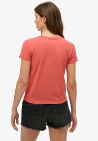 Kvinde set bagfra iført en koralfarvet T-shirt med korte ærmer og sorte denimshorts med frynser mod en hvid baggrund.