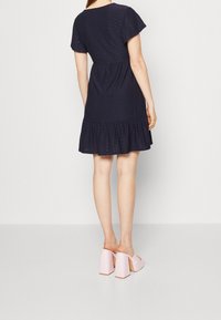 Frau in einem navyblauen Kurzarmkleid mit Rüschensaum, stehend mit dem Rücken zur Kamera, und hellrosa Plattformsandalen mit hohen Absätzen.