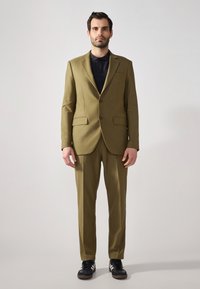 Completo verde oliva con giacca su misura dotata di revers a taglio, pantaloni abbinati e una camicia nera sottostante. Sono indossate sneakers nere.