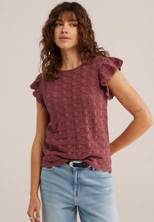 Naine, kellel on lokkis juuksed, seljas lilla heegeldatud lühikeste varrukatega top ja helesinised teksad, seisab ühe käega puusal, vaadates kõrvale.