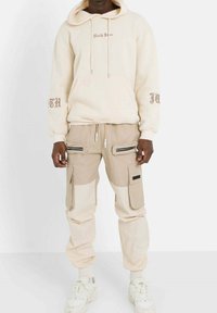 Beige hoodie med framfickan och skriptlogotyp, i kombination med ljusbruna cargo-byxor med flera dragkedjor och klafffickor. Vita sneakers.
