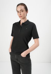 Černé polo tričko vyrobené z bavlny, s klasickým límečkem, krátkými rukávy, tříknoflíkovou légou a malým logem na hrudi.