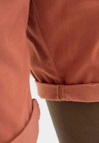 Rust-colored fabric with a smooth texture, folded hem, and visible stitching details. Close-up view of a garment's side section.