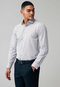 Camisa de botones en blanco con patrón a cuadros en azul y rojo, de manga larga, con cuello, combinada con pantalones oscuros y un cinturón negro.