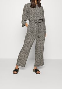 Combinaison à motifs noir et blanc avec ceinture à nouer, manches longues et coupe pantalon large, associée à des sandales noires sur un fond uni.