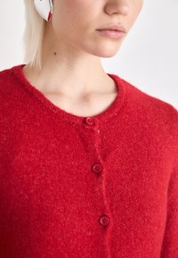 Cardigan de malha vermelha com um decote redondo, cinco botões vermelhos e uma textura suave. Vista aproximada da parte superior do corpo e da área do decote.