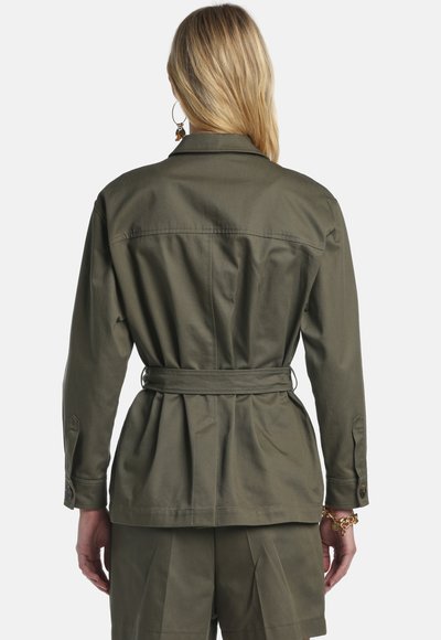 Woman with blonde hair wearing an olive green belted jacket and matching shorts, shown from the back against a plain background.