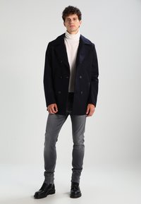 Cappotto doppiopetto blu scuro con bottoni grandi, indossato sopra un maglione a collo alto chiaro, abbinato a jeans grigi slim-fit e stivali neri stringati.