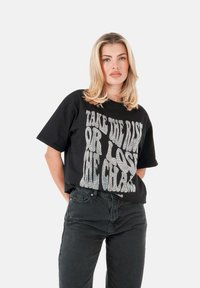 Camiseta de algodón negra con corte oversize, que presenta un gran texto gráfico blanco en la parte delantera. Combinada con jeans oscuros para un look casual.