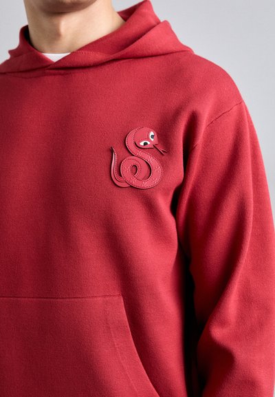 Felpa rossa con cappuccio e cordino, caratterizzata da un emblema di serpente testurizzato in rilievo dello stesso colore, con occhi e una posa giocosa. Tessuto morbido.