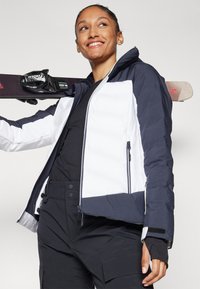 Skijacke mit gepolstertem Design in Weiß und Marineblau, ausgestattet mit Reißverschlüssen, Taschen und einer Kapuze, kombiniert mit schwarzen Skihosen.