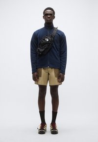 Ναυτικό φλις τζάκετ, χακί σορτς με τσέπες, μαύρη τσάντα χιαστί, μαύρες κάλτσες και ορειβατικά παπούτσια με κόκκινες λεπτομέρειες. Το μοντέλο στέκεται όρθιο.