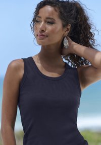 Top in tinta blu scuro a costine con scollatura rotonda. Il modello ha capelli ricci fino alle spalle ed indossa un orecchino a frangia. Sfondo da spiaggia.