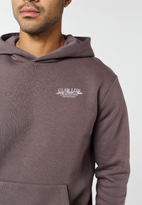 Férfi, mauve színű kapucnis pulóverben, amelynek mellkasán fehér felirattal szerepel: "CLSR LDN Luxury British Atelier Closure Design Studio Crafted in Soho".