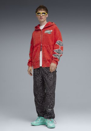 Giovane uomo con occhiali da sole gialli, felpa rossa con stampe grafiche, pantaloni neri con fantasia e sneakers turchesi, in piedi su sfondo grigio.