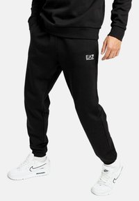 Pantaloni jogger neri realizzati in tessuto morbido, con cintura elastica, tasche laterali e un piccolo logo bianco sulla coscia.