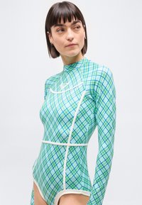 Γυναίκα με κοντά σκούρα μαλλιά που φοράει ένα μακρυμάνικο κορμάκι με πράσινο και μπλε καρό σχέδιο, κοιτάζει προς τα αριστερά της σε ένα απλό λευκό φόντο.