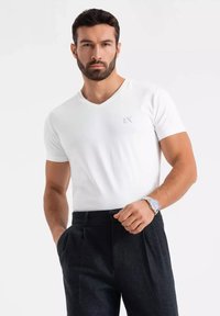 Biała koszulka z dekoltem w serek i krótkimi rękawami, wykonana z miękkiego materiału, z małym szarym logo na piersi, w połączeniu z ciemnymi eleganckimi spodniami.