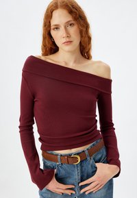 Bordeauxfarbenes, schulterfreies Oberteil aus geripptem Stoff mit langen Ärmeln und taillierter Passform, kombiniert mit blauen Jeans und einem braunen Gürtel.