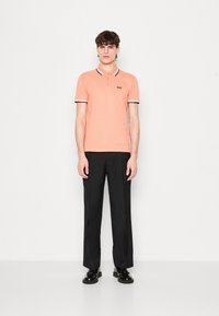 Poloshirt in Koralle mit schwarz-weißen gestreiften Kragen und Bündchen, kombiniert mit schwarzen Hosen und schwarzen Schuhen. Das Modell steht vor einem weißen Hintergrund.
