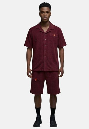 Homme portant une chemise bordeaux à manches courtes boutonnée assortie et un short, avec des chaussettes noires et des chaussures noires, debout devant un fond uni.