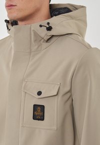 Giacca impermeabile beige con colletto alto, cappuccio regolabile, tasca frontale e accessori neri. Presenta una patch con logo sulla tasca.