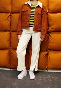 Oranžová sametová bunda s bílým beránkovým límcem, kombinovaná se zeleným a černým pruhovaným tričkem, béžovými rozšířenými kalhotami a levandulovými botami.