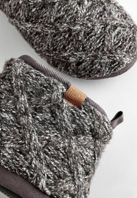 Gestrickter grau-weißer Hausschuh mit einem strukturierten Muster, versehen mit einem braunen Lederlogo-Akzent und einer weichen, gepolsterten Sohle.