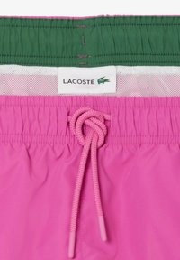 Plavky Lacoste s ružovým základom, zeleným pásom a šnúrkou na stiahnutie. Obsahujú sieťovú podšívku a značka je umiestnená na páse.