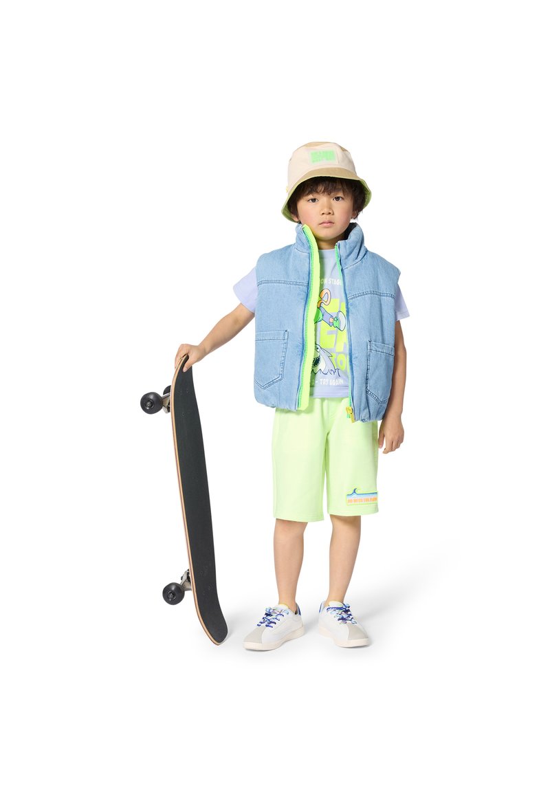 Τζιν γιλέκο με πράσινη επένδυση, μπλούζα με γραφικά, νεον-πράσινα σορτς και λευκά αθλητικά παπούτσια με μπλε κορδόνια, κρατώντας ένα μαύρο σκέιτμπορντ.