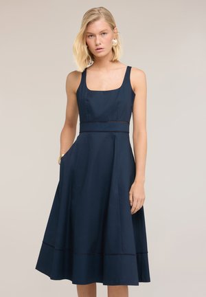 Blonde vrouw in een mouwloze marineblauwe midi-jurk met een vierkante halslijn, staand met één hand in haar zak tegen een neutrale achtergrond.