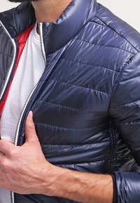 Giubbotto piumino blu navy con finitura lucida, motivo a righe orizzontali, colletto rialzato e chiusura con zip. Fodera rossa visibile all'interno.