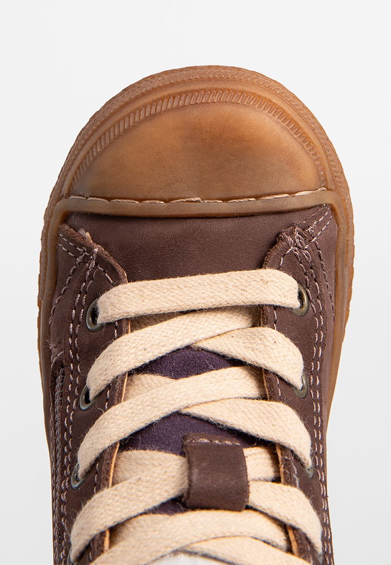 Scarpa in pelle marrone con lacci beige incrociati e punta in gomma rotonda, vista dall'alto in primo piano su uno sfondo bianco.