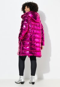 Chaqueta puffer metálica fucsia con capucha, con diseño acolchado, cremalleras laterales y corte a media longitud, combinada con jeans negros y botas plateadas.